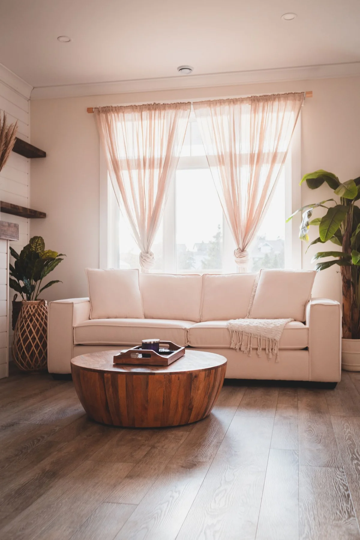 Cream sofa beneath a curtained window, with a round wooden coffee table and plants